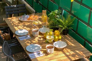 Una mesa de madera dispuesta con platos, vasos y una botella, rodeada de plantas y una cerca verde.