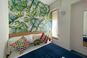 A bedroom with a bed, colorful pillows, a tropical leaf wallpaper, a window with curtains, and a nightstand with a lamp.