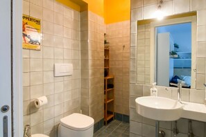 Un baño con inodoro, lavabo, ducha y estante con artículos de aseo.