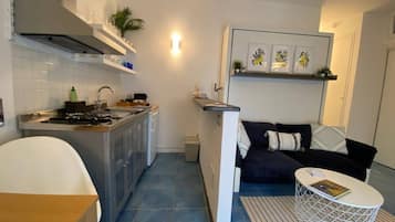 Ett kompakt kök med spis, diskho och skåp, en matplats med bord och stolar samt ett vardagsrum med soffa och soffbord.