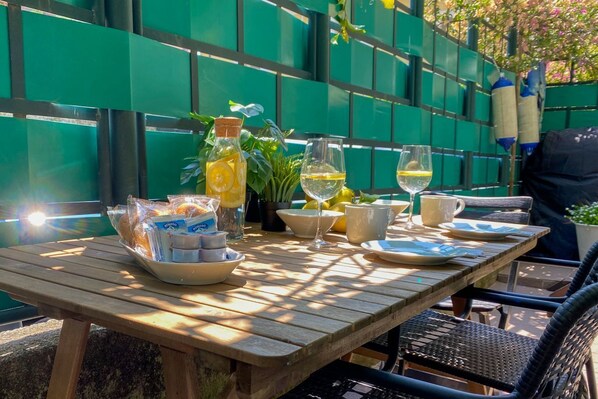 A wooden table with a breakfast setup including bread, butter, and beverages, set against a green fence.