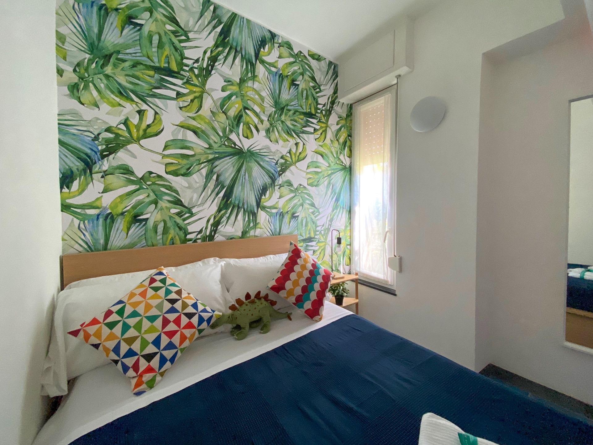 Une chambre à coucher avec un lit, des oreillers, une fenêtre et un mur orné d’un motif de feuilles tropicales.