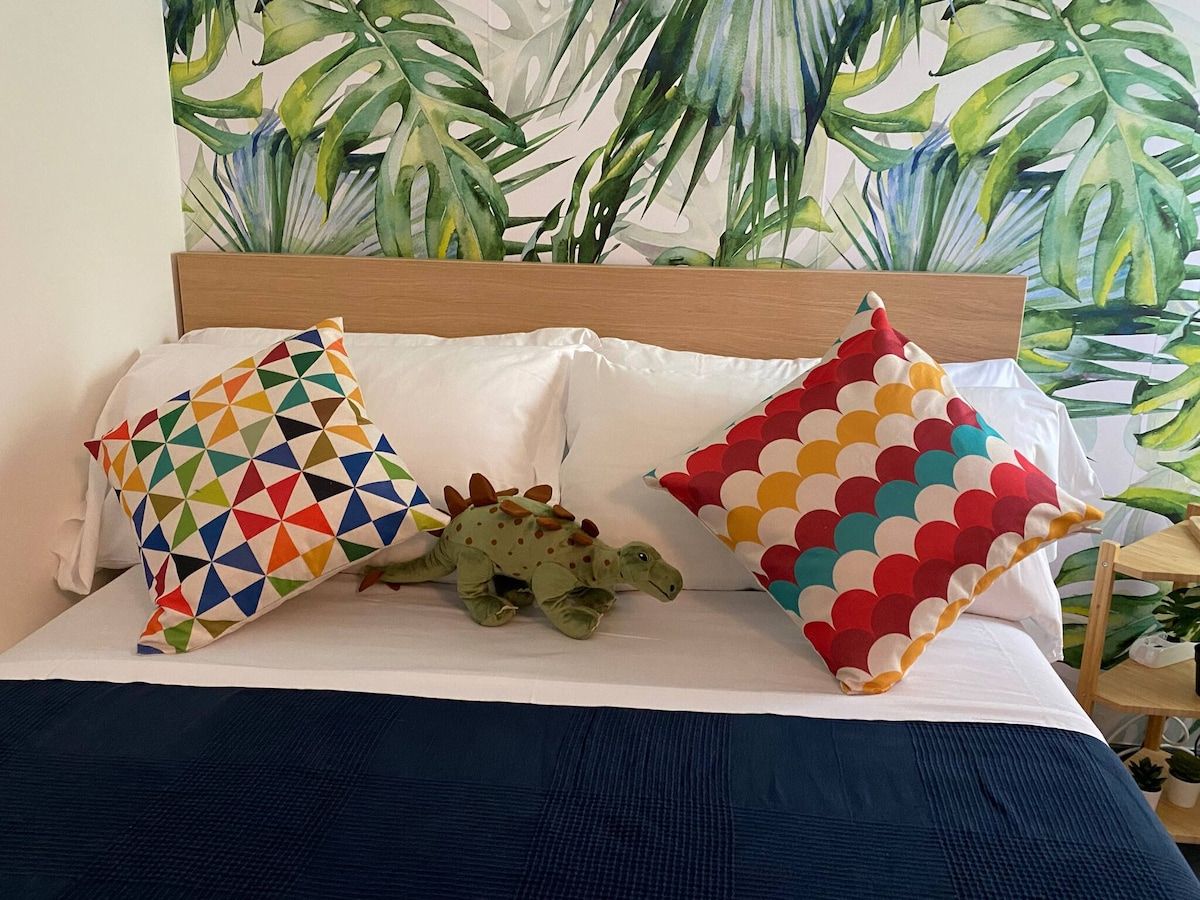 Ein Bett mit einem Holz-Kopfteil, zwei Kissen mit geometrischen Mustern und ein grünes Dinosaurier-Spielzeug.