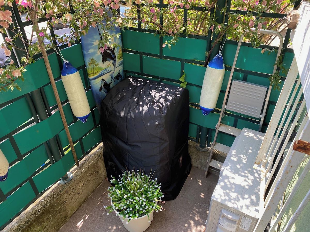 A covered outdoor seating area with a green fence, a covered trash bin, and a potted plant.