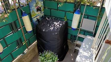 A covered outdoor seating area with a green fence, a covered trash bin, and a potted plant.