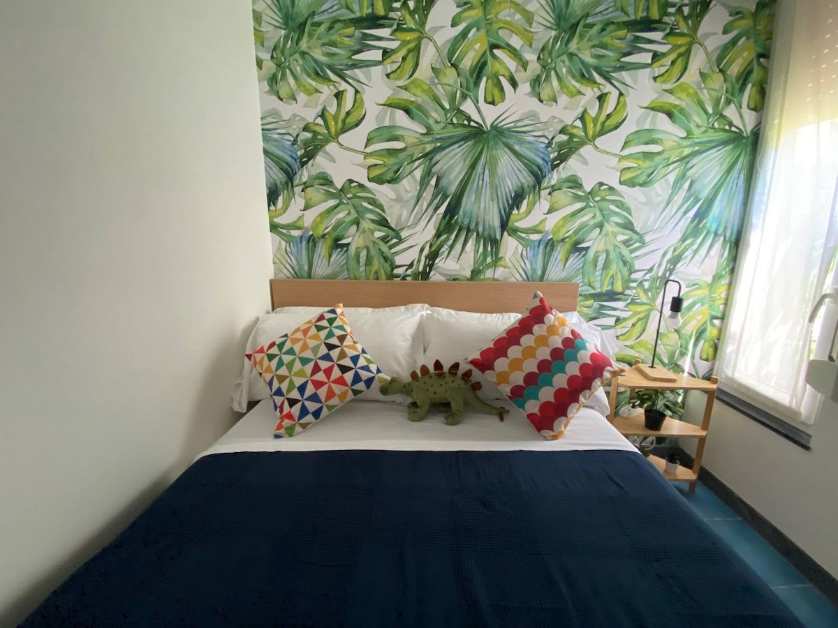 Un lit recouvert d’une couverture bleu foncé, deux oreillers ornés de motifs géométriques colorés et un jouet en forme de dinosaure posé sur le lit.