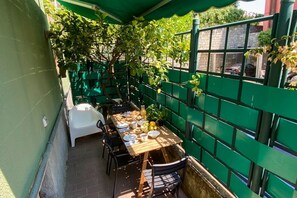 Un espacio comedor al aire libre, cubierto, con una mesa dispuesta para dos personas, rodeada de sillas y una cerca verde que delimita el área.