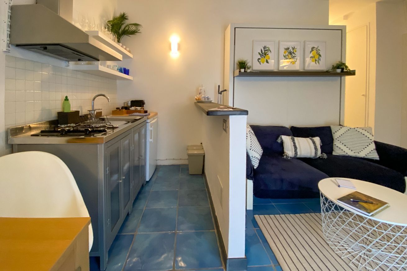 A compact kitchen with a stove, sink, and cabinets, a small trash bin, a sofa with patterned cushions, and a white coffee table.