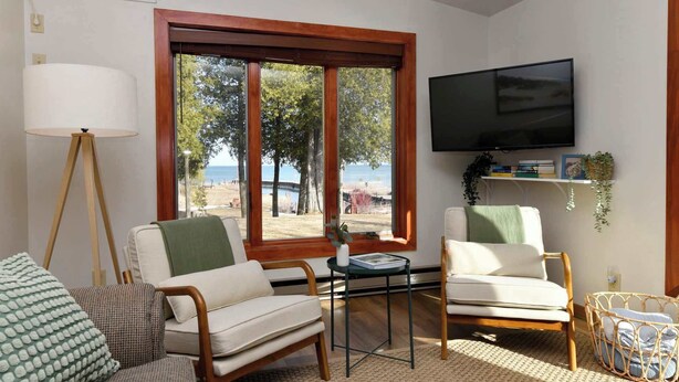 A living room with two chairs, a table, a TV, and a large window with a view of the beach.