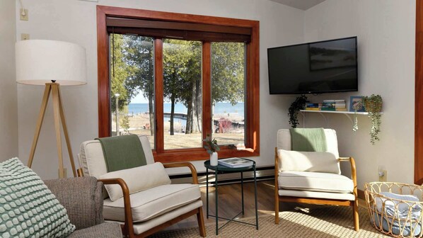 A living room with two chairs, a table, a TV, and a large window with a view of the beach.