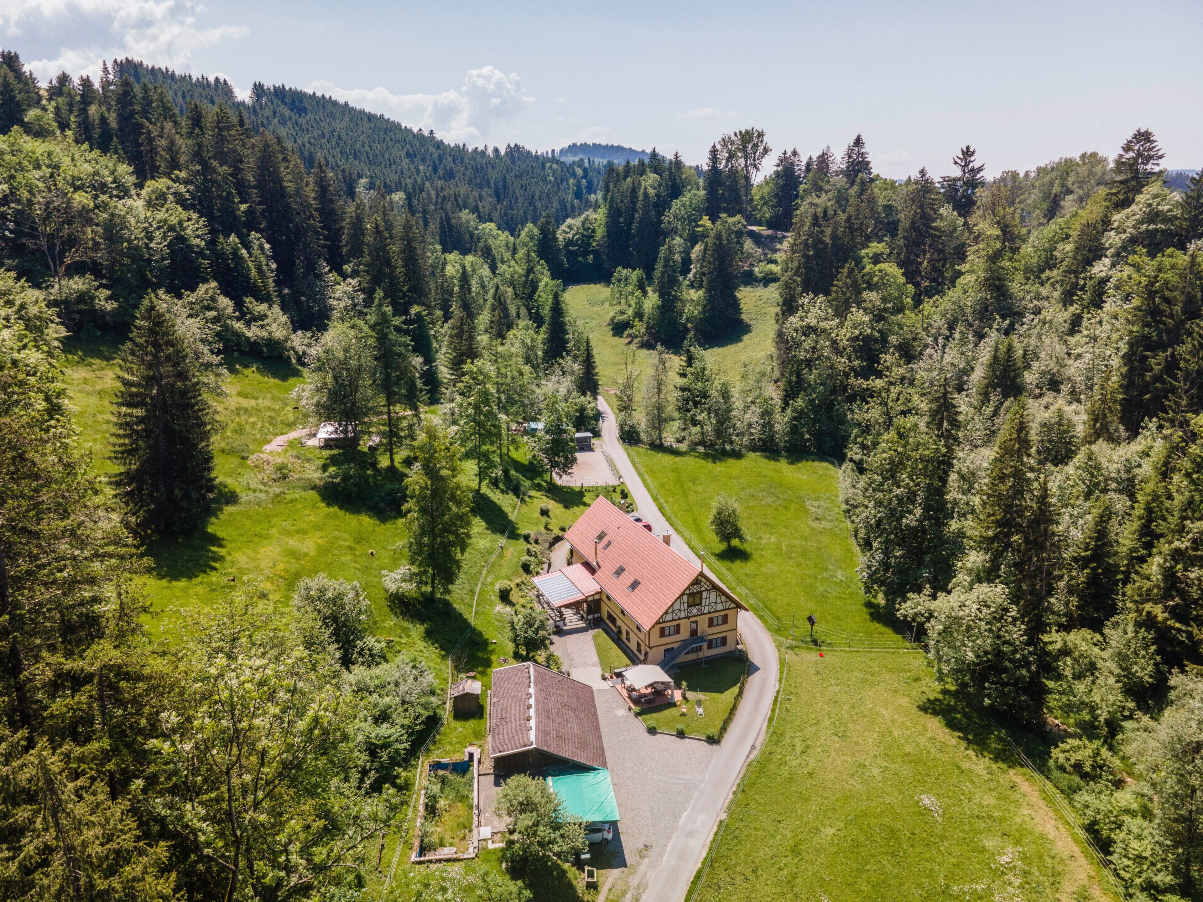 Une maison de campagne avec un toit rouge, entourée de champs verdoyants et d’une forêt dense.