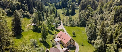 A rural house with a red roof, surrounded by lush green fields and dense forest.