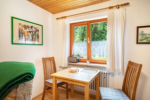 Une pièce avec une table en bois, deux chaises, un radiateur, une fenêtre avec des rideaux, un tableau encadré au mur et une couverture verte sur une chaise.