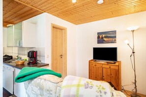 Une pièce comprenant un lit, un téléviseur posé sur un meuble en bois, un coin cuisine avec une machine à café et un plafond en bois.