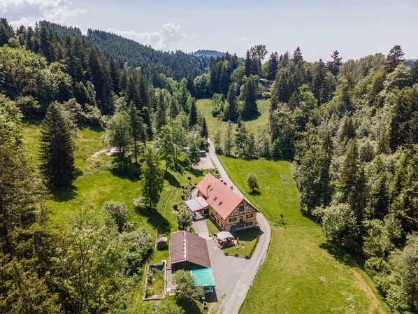 A large house with a red roof, surrounded by a lush green landscape and dense forest.