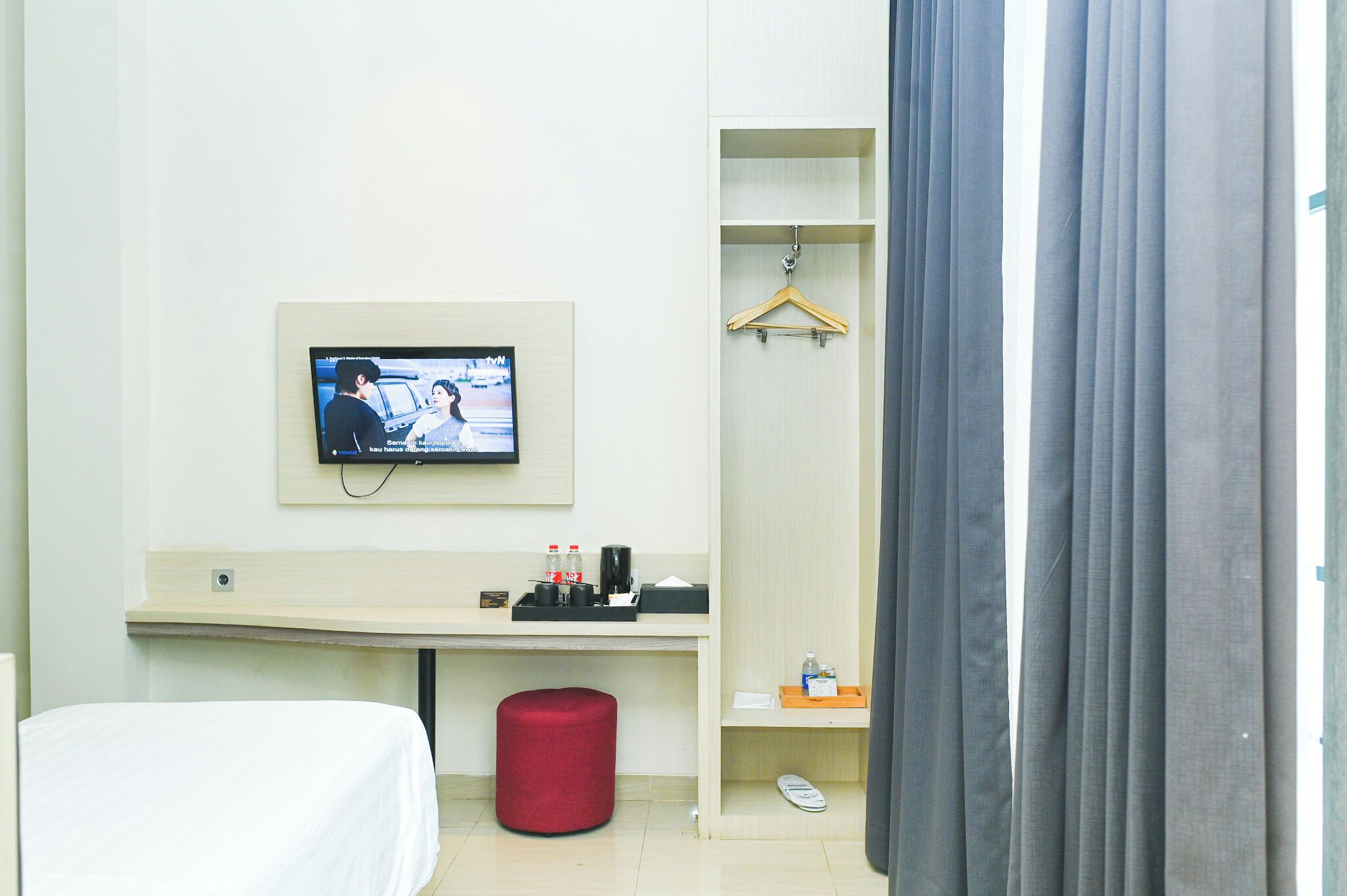Camera d'albergo con un letto, una scrivania con televisore, un poggiapiedi rosso, un cappotto appeso e uno scaffale per scarpe.