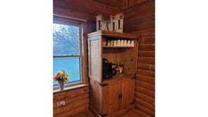 En stuga med träpanel på insidan, ett fönster, ett skåp med hyllor och en vas med blommor.
