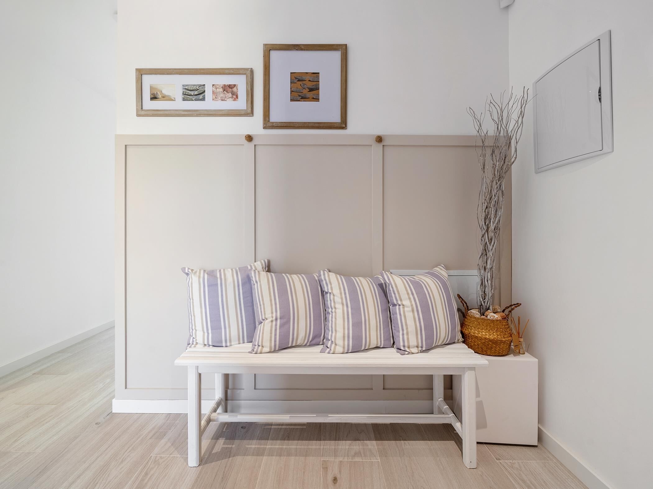 A white bench with striped cushions, a woven basket with decorative items, and framed artwork on the wall.