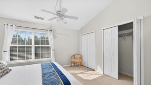 Une chambre à coucher comprenant un lit, un ventilateur de plafond, une fenêtre avec des stores, une chaise et un placard.