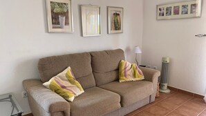 A living room with a brown sofa, two cushions, a glass coffee table, a floor lamp, and a standing heater.