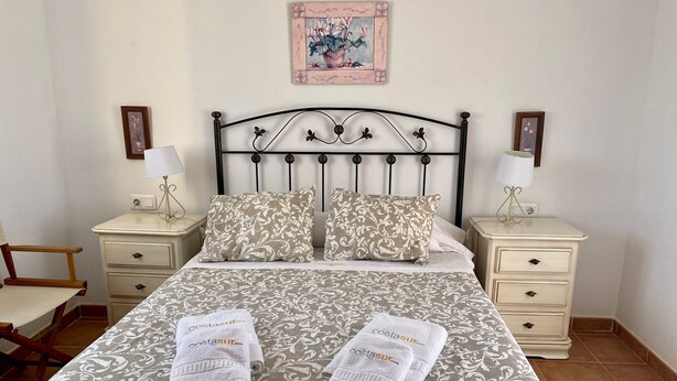 A neatly made bed with a patterned bedspread, two pillows, and two folded towels on the bed. There are two bedside tables with lamps and framed pictures on the wall above.