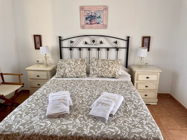 A neatly made bed with a patterned bedspread, two pillows, and two folded towels on the bed. There are two bedside tables with lamps and framed pictures on the wall above.