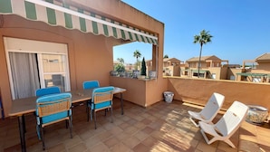 A terrace with a table and chairs, a covered area with a striped awning, and a view of neighboring houses and palm trees.