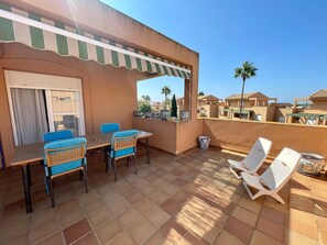 A terrace with a table and chairs, a covered area with a striped awning, and a view of neighboring houses and palm trees.