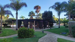 A covered outdoor area with palm trees, a brick pathway, and a building in the background. - Prado Golf Apartment with shared swimming pool (Ayamonte)