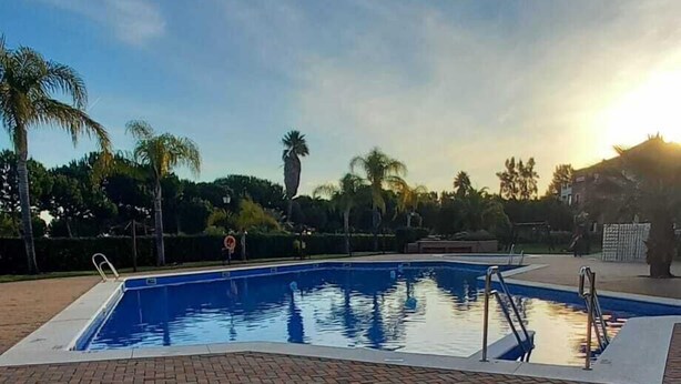 A swimming pool with a brick patio area, surrounded by palm trees and a clear sky. - Prado Golf Apartment with shared swimming pool (Ayamonte)