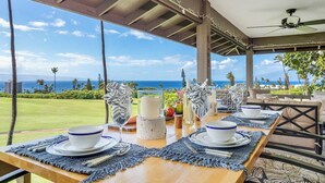 Un coin repas avec vue sur l’océan, une table en bois dressée avec de l’assiette blanche et des serviettes bleues, et un ventilateur de plafond.