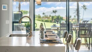 Une cuisine dotée d’un grand îlot central, d’un évier et offrant une vue sur un paysage luxuriant grâce à de grandes fenêtres.