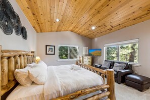 A bedroom with a wooden headboard, a bed with white linens, a leather sofa, a television, and a window with a view of trees.