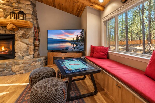 A cozy room with a stone fireplace, a television, a game table, and a window with a view of trees.