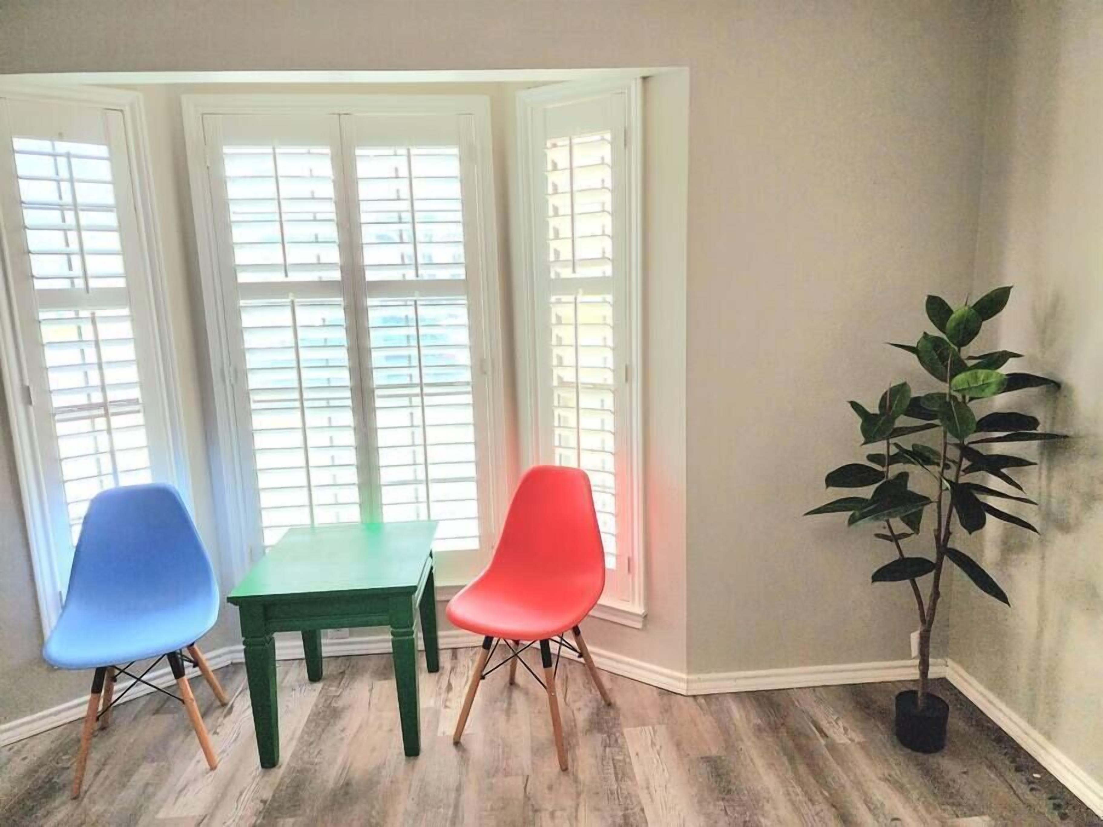 Ein Zimmer mit Tisch, zwei Stühlen, einer Topfpflanze und einem Fenster mit Jalousien.
