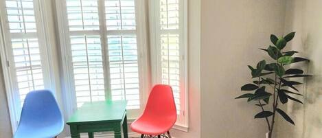 Ein Zimmer mit Tisch, zwei Stühlen, einer Topfpflanze und einem Fenster mit Jalousien.