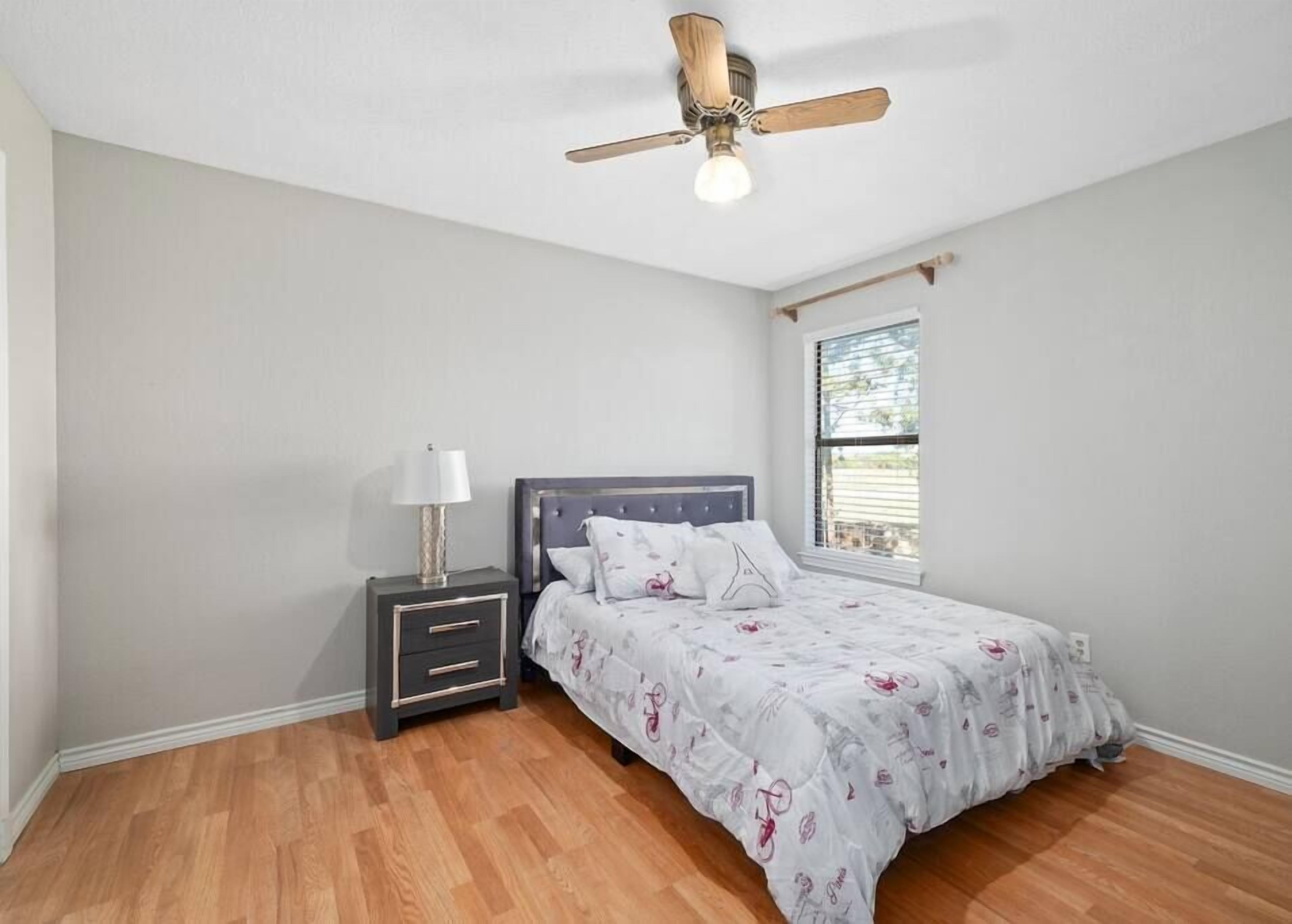 A bedroom with a bed, nightstand, lamp, ceiling fan, and a window.