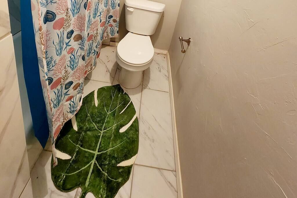 Une salle de bain avec des toilettes, un rideau de douche orné de motifs représentant la vie marine, et un grand tapis vert en forme de feuille sur le sol.
