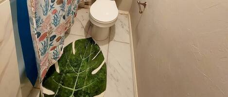 Une salle de bain avec des toilettes, un rideau de douche orné de motifs représentant la vie marine, et un grand tapis vert en forme de feuille sur le sol.