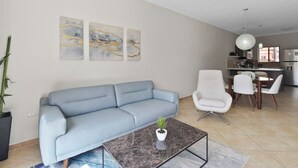 Una sala de estar moderna con un sofá azul claro, una mesa de centro con una planta, y un comedor con sillas blancas y una cocina al fondo.