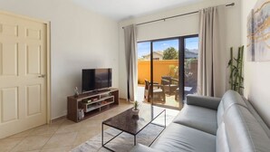 Una sala de estar con un sofá, un televisor sobre un mueble de madera, una mesa de centro y un ventanal corredero que da a un patio exterior con una mesa y sillas.