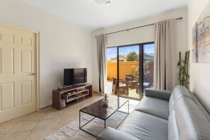 Uma sala de estar com sofá, TV sobre um suporte de madeira, mesa de centro e porta de vidro deslizante que dá para um pátio externo com mesa e cadeiras.