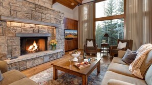 Un salon chaleureux avec une cheminée en pierre, des fauteuils confortables et une grande fenêtre donnant sur la forêt.