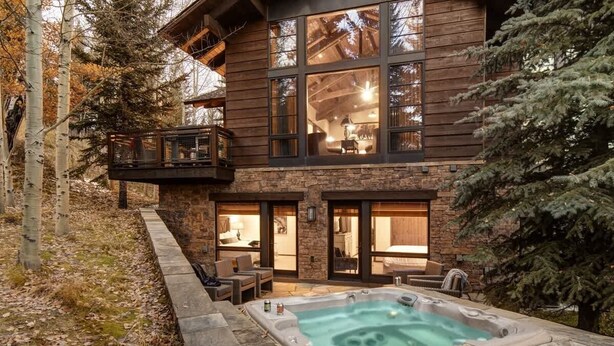 Une cabane en rondins à deux étages, avec des fondations en pierre, un jacuzzi et un balcon donnant sur une zone boisée.