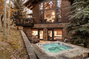 A two-story log cabin with a stone foundation, a hot tub, and a balcony overlooking a forested area.