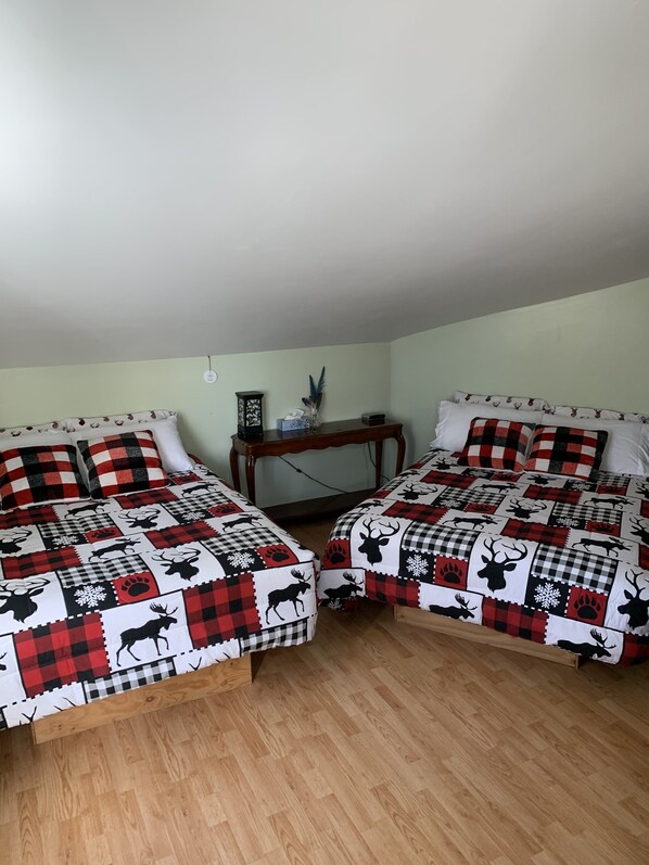 A bedroom with two beds, each with a checkered bedspread featuring animal prints. There is a wooden nightstand between the beds with a lamp and a decorative item. - Auberge de Ferme-Neuve (Ferme-Neuve)