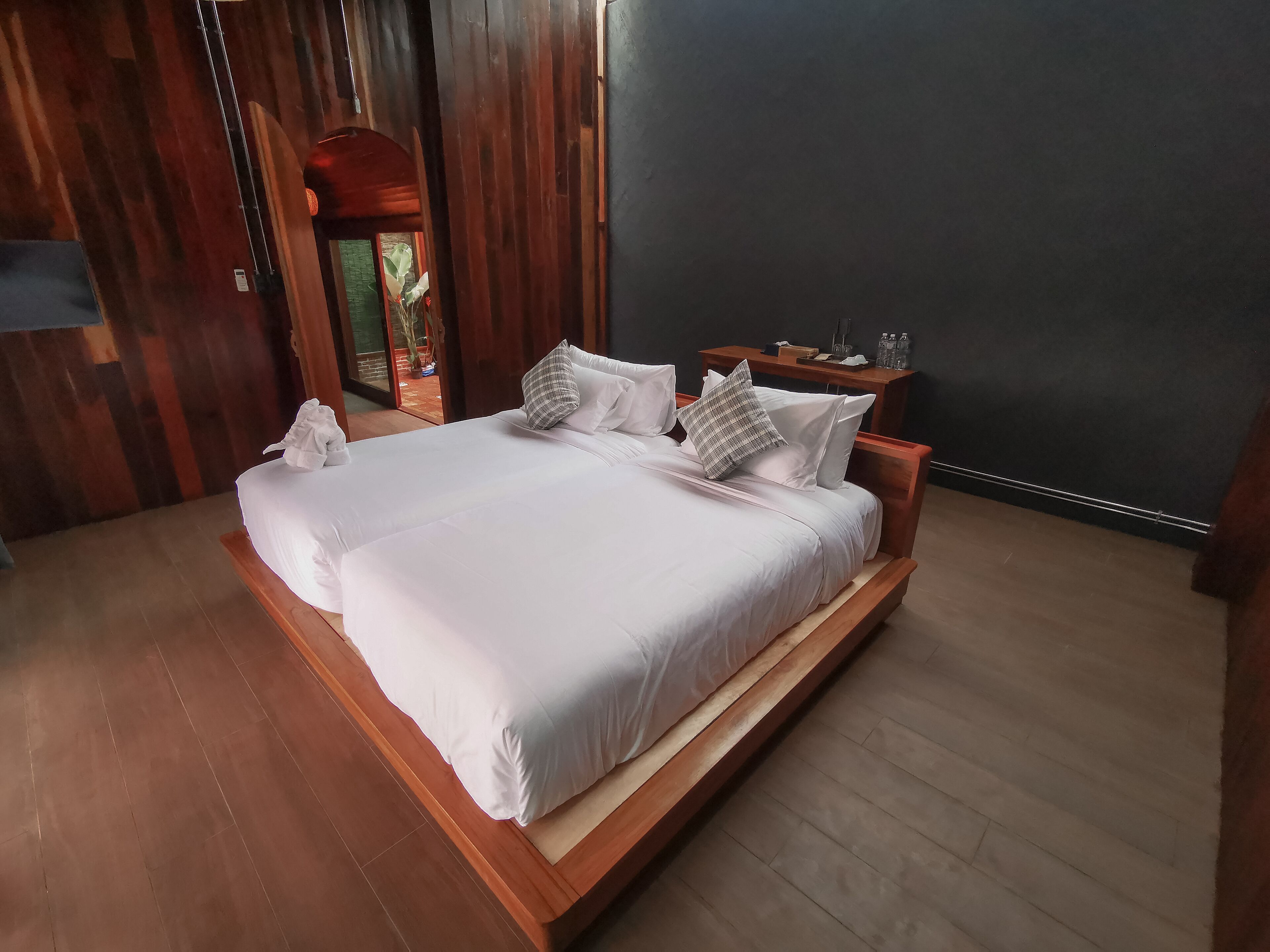 Een groot bed met witte lakens en meerdere kussens, een houten nachtkastje met een lamp, en een donkere muur.