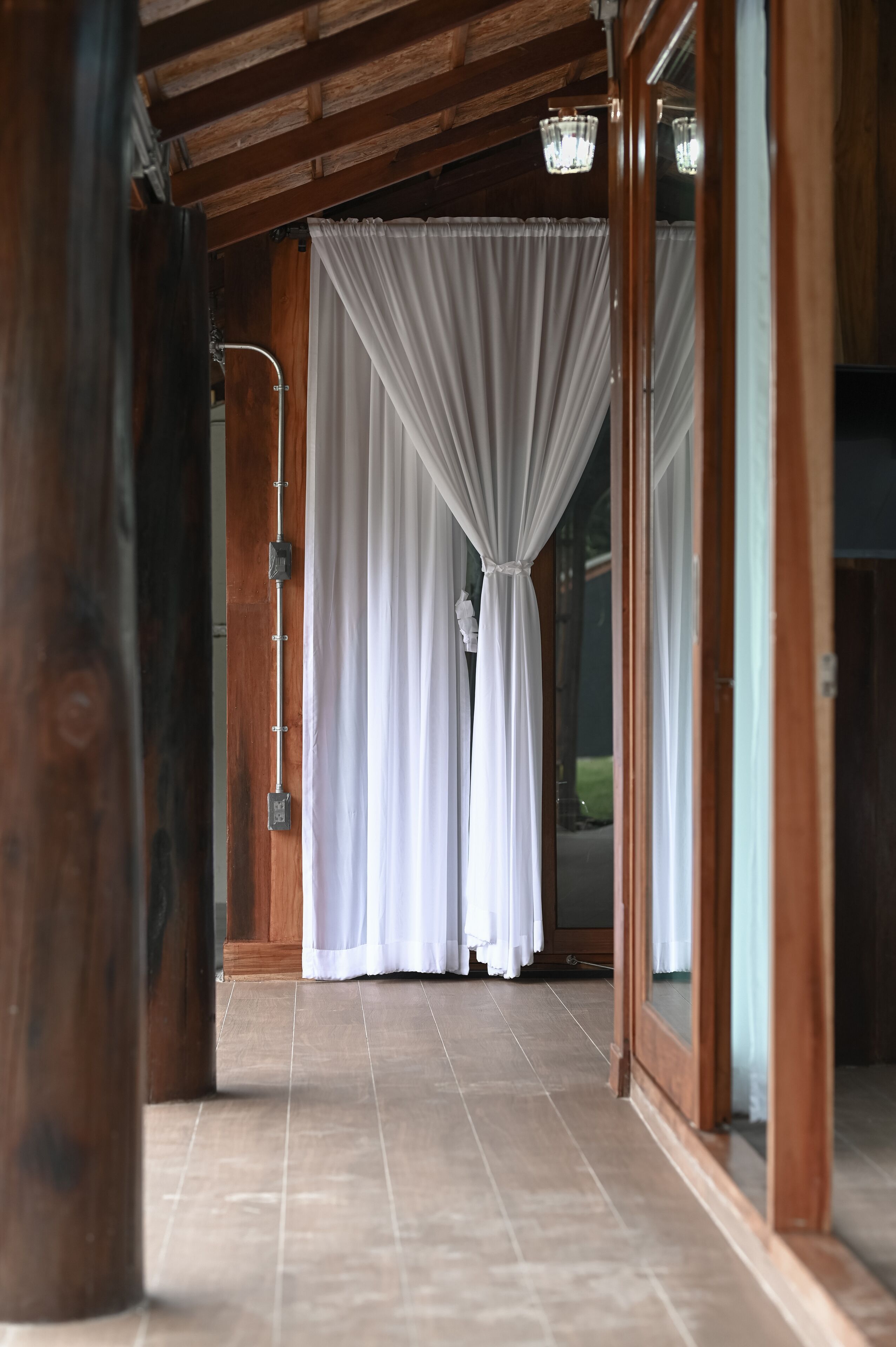 A corridor with wooden beams, white curtains, and a glass door leading outside.