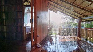 Una terraza de madera con barandilla metálica, una puerta de madera que da a una habitación con cama y vistas al verdor exterior.