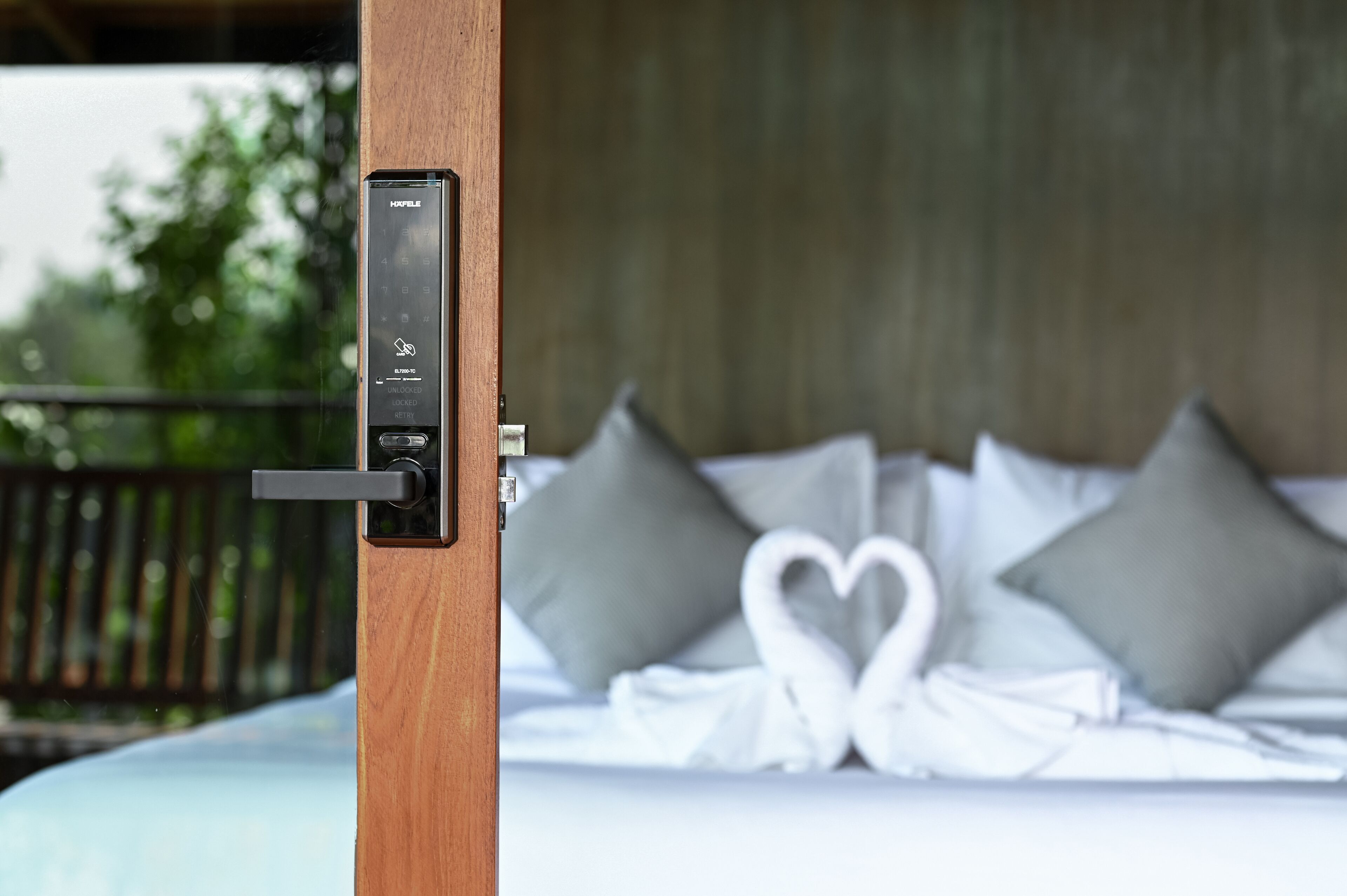 A hotel room with a bed featuring a heart-shaped decoration, a control panel on the door, and a view of greenery outside.